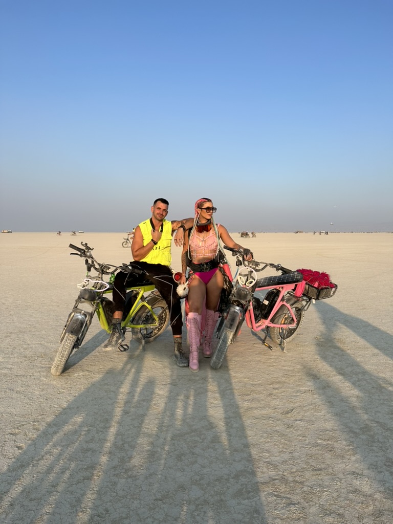 Two DUST riders on neon green and pink eBikes on the open playa